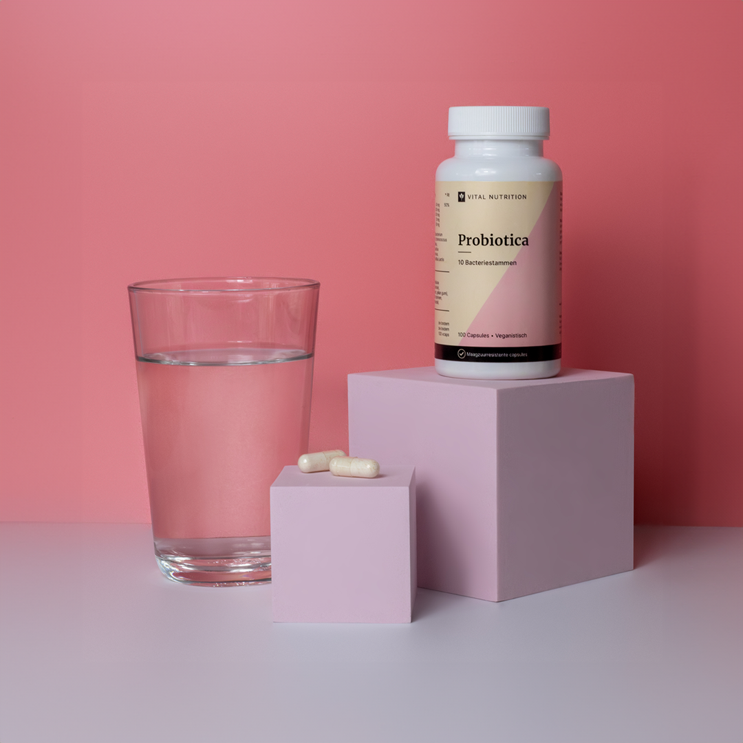 Vital Nutrition Probiotica op verhoging met roze achtergrond, glas water en capsules zichtbaar