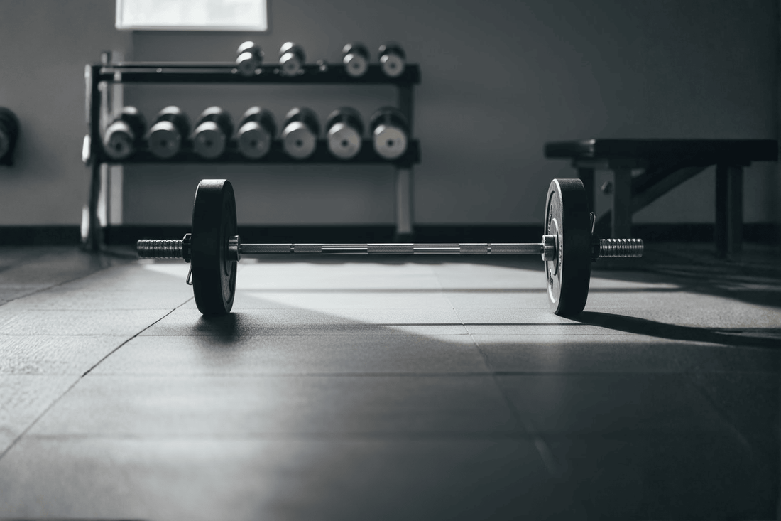 Close-up van halterstang met zwarte schijven op een lichte sportvloer in natuurlijk daglicht.